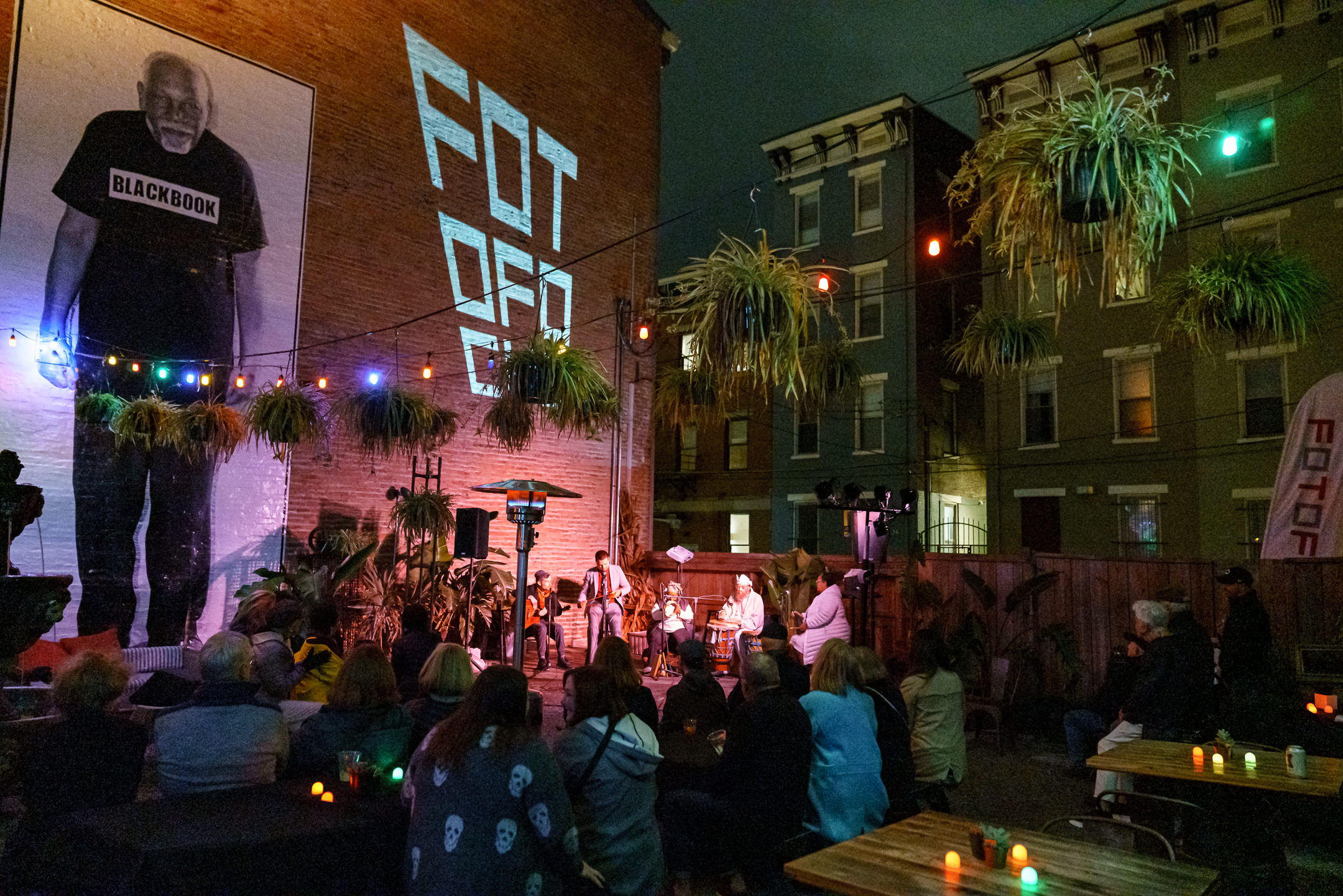 Musical performers and audience in an outdoor patio surrounded by city buildings